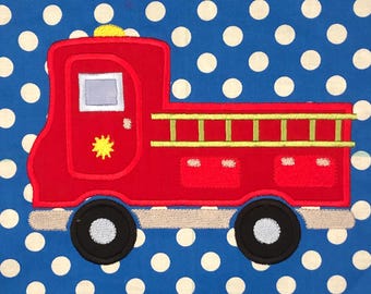 May include: A red fire truck applique on a blue fabric background with white polka dots. The truck features a yellow light on top, a grey window, a yellow ladder on the side, and black wheels with grey centers. The text JJ's Appliques is at the bottom.