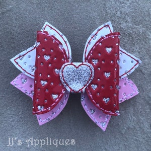 Op de afbeelding: Een gelaagde strik in rood, roze en wit met een zilveren glitterhart in het midden. De strik is versierd met kleine witte hartjes.