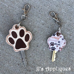 May include: Two keychains with paw print designs. One keychain is a brown paw print on a white background with a red stitch. The other keychain is a white background with brown paw prints and bones and the word "woof" in brown letters.