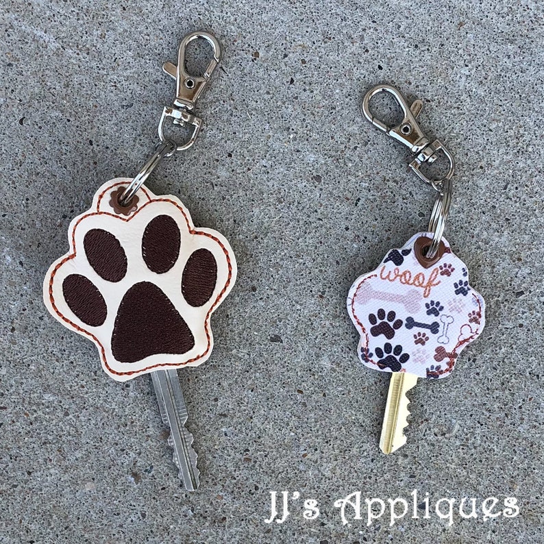 May include: Two keychains with paw print designs. One keychain is a brown paw print on a white background with a red stitch. The other keychain is a white background with brown paw prints and bones and the word "woof" in brown letters.