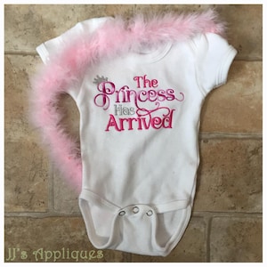 May include: White baby onesie with pink and silver embroidery that reads "The Princess Has Arrived".