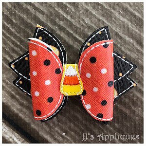 Op de afbeelding: Een zwarte en oranje gepolka-dotte strik met een gele snoepmais-applicatie. De strik is gestikt met wit garen en heeft een gegolfde rand.