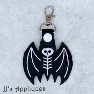 May include: A black bat-shaped keychain featuring a white embroidered skeleton design. The bat is attached to a silver-toned metal lobster clasp by a black strap secured with a white snap button. The text JJ's Appliques is visible at the bottom.