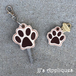 May include: Two brown and white paw print keychains with red stitching. The larger keychain has a silver key ring and a key attached. The smaller keychain has a gold key ring and a key attached. The text "JJ's Appliques" is visible at the bottom of the image.