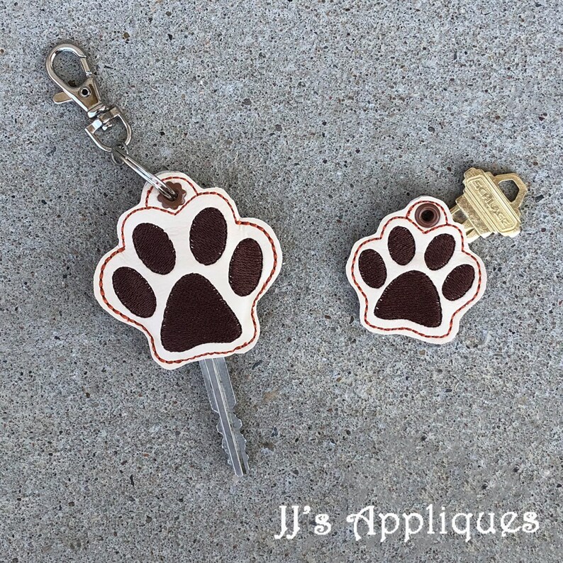 May include: Two brown and white paw print keychains with red stitching. The larger keychain has a silver key ring and a key attached. The smaller keychain has a gold key ring and a key attached. The text "JJ's Appliques" is visible at the bottom of the image.