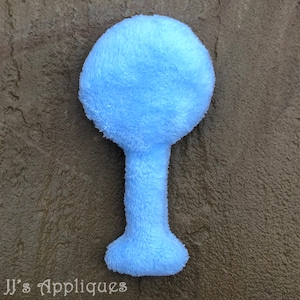 以下が含まれることがあります： ライトブルーの、ぬいぐるみのような木の形をしたアップリケ。アップリケは丸みを帯びた上部、細い茎、フレアベースを持っています。テクスチャは柔らかくふわふわに見えます。「Jj's Appliques」の文字が下部にあります。