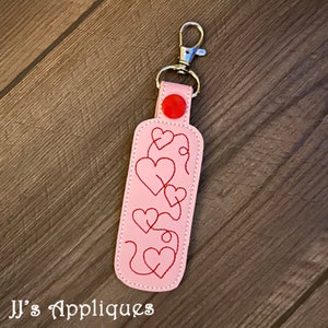 May include: Pink leather keychain with a red button and a silver clip. The keychain has a design of red hearts connected by a line.