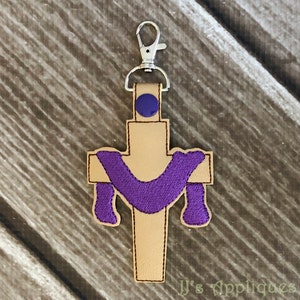 May include: A purple fabric cross with a beige background and a silver clip. The cross is decorated with a purple fabric drape.