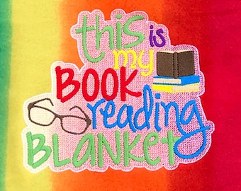 May include: A colorful embroidered patch with the text "This is my book reading blanket" and an image of glasses and a stack of books. The patch is on a rainbow-colored background.