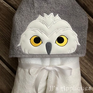 May include: A gray hooded towel with a white embroidered owl face. The owl has yellow eyes and a gray beak. The towel has a white ribbon tied around it.