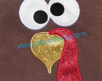 May include: A brown fabric background featuring a turkey face applique. The design includes two large white eyes with black pupils, a gold glitter heart-shaped beak, and a red glitter wattle. The text www.jjsappliques.com is printed in blue.