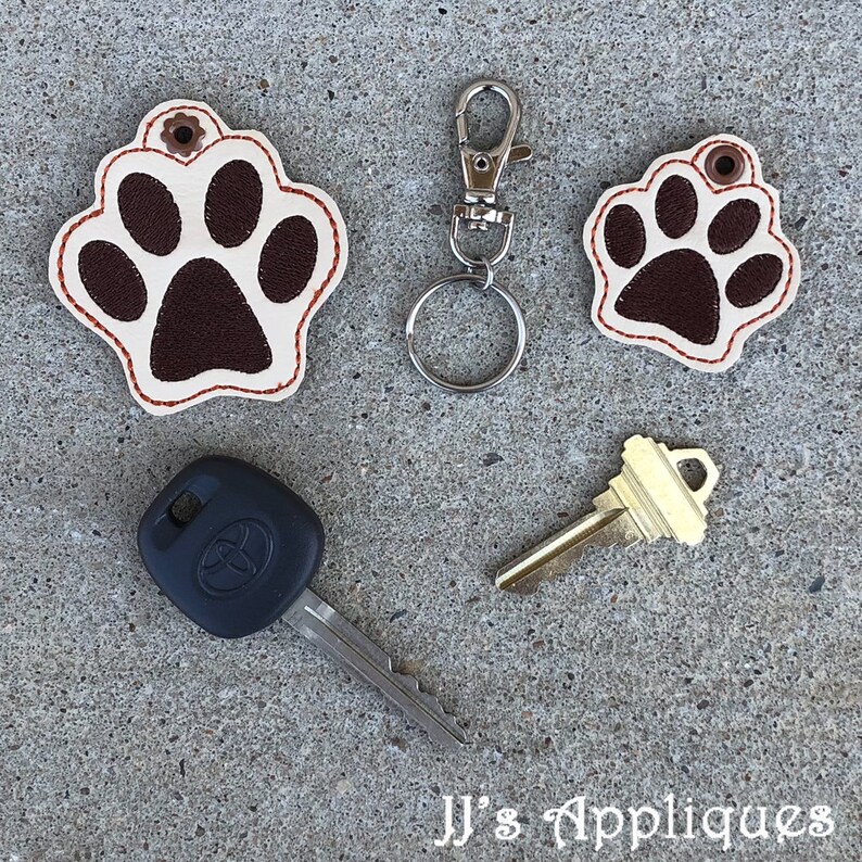 May include: Two brown leather paw print keychains with red stitching. The larger keychain is attached to a silver key ring. A black car key and a gold house key are also pictured. The image includes the text "JJ's Appliques".