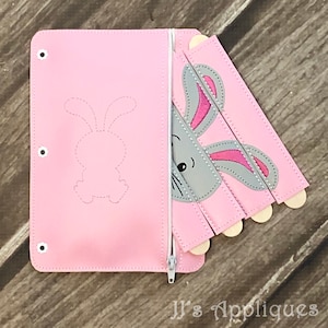 May include: A pink felt zippered pouch with a gray bunny face peeking out from behind the flap. The bunny face is made of felt and has pink ears and a white nose. The pouch has a white outline of a bunny on the front.