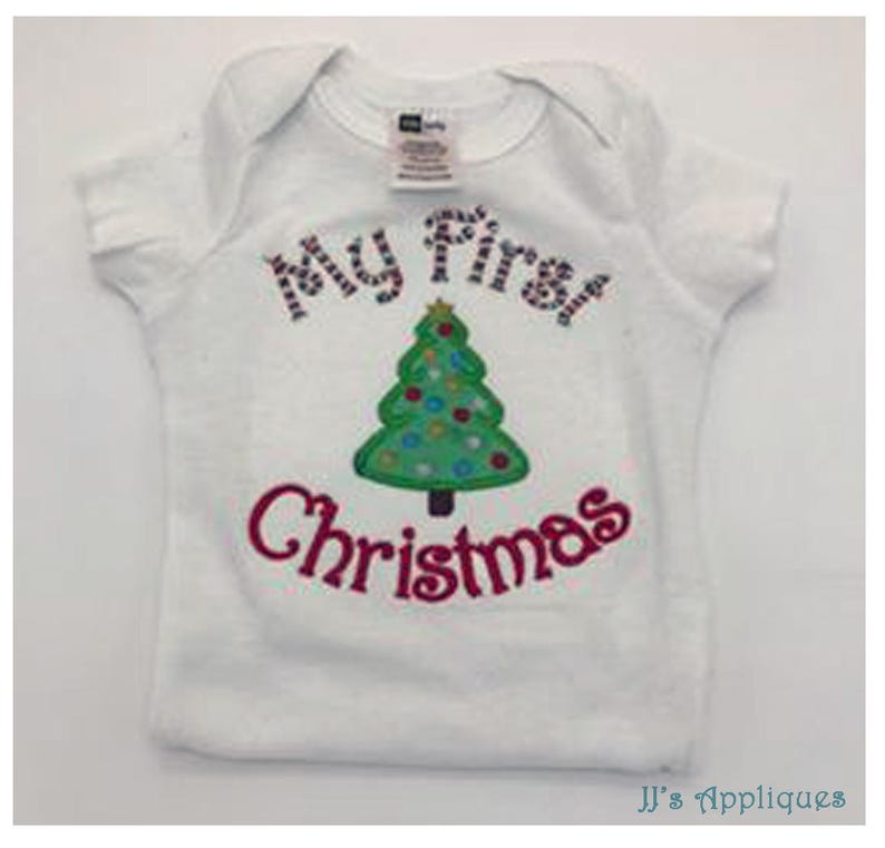 May include: A white short-sleeved baby bodysuit featuring an embroidered green Christmas tree with colorful circular ornaments. The text "My First" is stitched in a red and white striped pattern above the tree, and "Christmas" in red below.