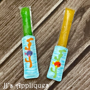 May include: Two popsicle holders with a blue background and a sea-themed design. One holder has a green popsicle and a blue fish with pink fins. The other holder has a yellow popsicle and an orange fish with red fins. Both holders have a green seaweed design.