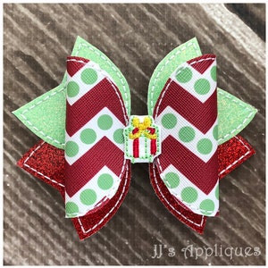 Op de afbeelding: Een rode en groene kerst strik met witte stippen en een chevron patroon. De strik heeft een klein groen en rood cadeaudoosje met een gele strik in het midden.