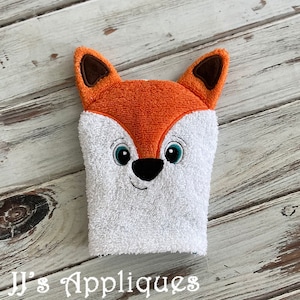 May include: A white washcloth with a fox face embroidered on it. The fox has orange fur, brown ears, and blue eyes.
