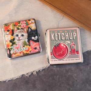 Könnte beinhalten: Zwei Zigarettenetuis aus Metall. Eines zeigt ein Blumendesign mit mehreren Katzen und dem Namen "Fisher". Das andere Etui hat ein Vintage-Ketchup-Etikettendesign mit einer Tomatenillustration und dem Text "Tomato Ketchup".