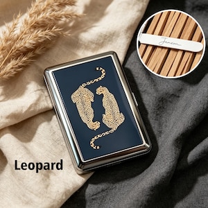 Könnte beinhalten: Ein rechteckiges Zigarettenetui aus Metall mit einer dunkelblauen Frontplatte, die zwei goldfarbene Leoparden-Illustrationen zeigt. Das Innere, in einem kreisförmigen Ausschnitt zu sehen, enthält Holztrennwände und einen weißen Metallsteg mit der Gravur Jenson.