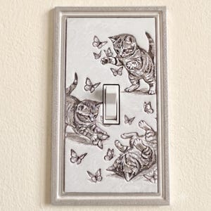May include: A rectangular light switch cover plate featuring a raised, monochromatic relief design of three kittens playing with several butterflies. The plate has a silver-toned border and a central white toggle switch mechanism.