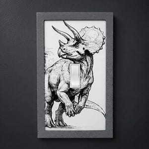 May include: A rectangular light switch cover featuring a black and white line drawing of a triceratops dinosaur. The cover has a textured dark gray border and a central white rocker switch. The dinosaur illustration covers the entire plate surface.