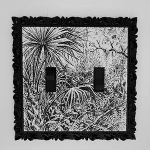 May include: A double light switch cover featuring a black and white botanical illustration of palm fronds and foliage. The plate is encased in a decorative black frame with a carved, ornate border and includes two black rocker switches.