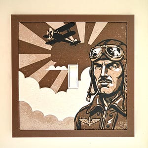May include: A square light switch cover featuring a brown and white graphic illustration of a pilot wearing goggles and a leather cap. The design includes a biplane flying in front of a sunburst pattern and clouds, framed by a solid brown border.