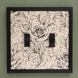 May include: A square double light switch plate featuring a black frame and a cream-colored center panel with a detailed black ink illustration of roses and swirling vines. Two vertical black rocker switches are centered on the plate.