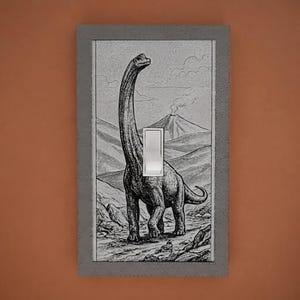 May include: A rectangular light switch cover featuring a black and white illustration of a long-necked dinosaur standing in a prehistoric landscape with a volcano in the background. The cover has a textured gray border and a central white rocker switch.