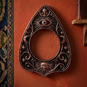 May include: A teardrop-shaped wall plaque featuring a central circular cutout. The dark, metallic-finished surface is decorated with an embossed eye at the top, a sun face at the bottom, and crescent moons and stars integrated into the scrollwork.
