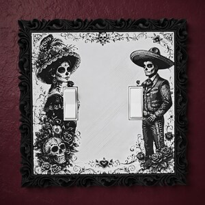 May include: A double light switch cover featuring a black and white illustration of a calavera couple in traditional attire, surrounded by roses and skulls. The plate is framed by a decorative black border and includes two white rocker switches.