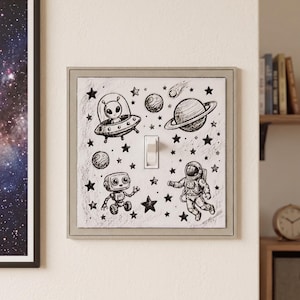 May include: A square light switch cover featuring a white background with black ink-style illustrations of an alien in a flying saucer, a planet with rings, a small robot, an astronaut, a comet, and various stars surrounding a central toggle switch.