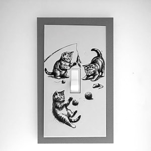May include: A rectangular light switch cover plate featuring a gray speckled border and a white center panel. The white panel displays three black and white illustrations of kittens playing with toys, including a fishing wand, a ball, and a mouse.