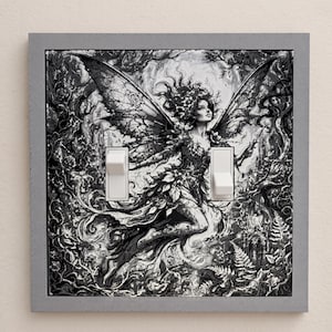 May include: A double light switch cover featuring a black and white illustration of a winged fairy surrounded by intricate forest foliage and vines. The rectangular plate has a gray border and two white toggle switches positioned in the center.