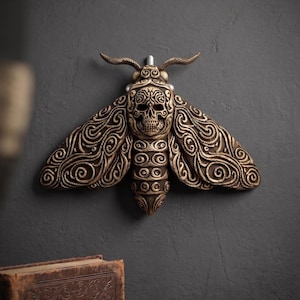 May include: A bronze-colored moth wall sculpture with intricate swirling patterns on the wings. The moth's body features a skull design, adding a gothic or macabre aesthetic. The sculpture is mounted on a dark gray wall.