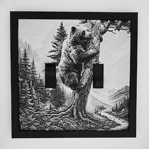 May include: A square double light switch cover featuring a black and white illustration of a bear climbing a tree in a forest landscape. The design includes two black toggle switches integrated into the scene, surrounded by a solid black frame.