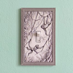 May include: A rectangular light switch cover plate featuring a detailed, raised relief design of two monkeys climbing through jungle vines and foliage. The plate has a textured, greyish-white finish with a central white toggle switch.