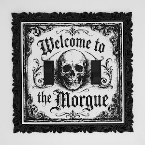 May include: A square double light switch cover featuring a black ornate frame and a white center plate. The center plate displays a black graphic of a human skull surrounded by scrollwork and the text Welcome to the Morgue in gothic font.