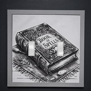 May include: A double light switch cover plate featuring a black and white illustration of a book titled Book of Spells. The artwork depicts an ornate, leather-bound tome resting on a fringed cloth, accompanied by a small sprig and a crystal.