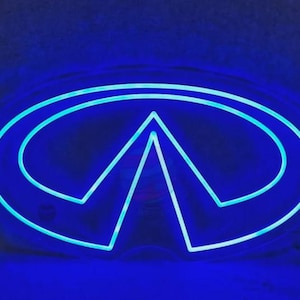 May include: A bright blue neon sign shaped like the Infiniti automotive logo. The design features a central triangular peak enclosed within an oval outline, emitting a vibrant, glowing light that illuminates the surrounding area in a deep blue hue.
