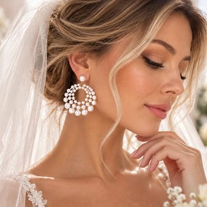 May include: A pair of circular drop earrings featuring multiple white pearls arranged in a concentric, tiered pattern. The design consists of a single pearl stud at the top, connected to a larger, open-work hoop structure adorned with smaller pearls.