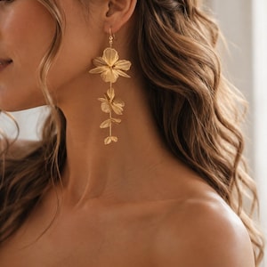 May include: A long gold-toned earring featuring a series of three delicate, layered floral shapes connected by a thin metal stem. The design hangs vertically from a simple hook, showcasing textured petals with a metallic finish and a slender structure.