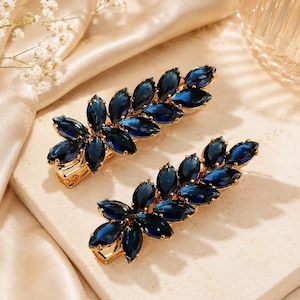 May include: Two hair clips featuring a gold-tone metal base adorned with marquise-cut dark blue crystals arranged in a leaf-like pattern. Each accessory has a secure metal clasp mechanism on the underside for attachment to hair strands.