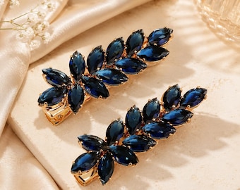 May include: Two hair clips featuring a gold-tone metal base adorned with marquise-cut dark blue crystals arranged in a leaf-like pattern. Each accessory has a secure metal clasp mechanism on the underside for attachment to hair strands.