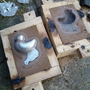 May include: Two wooden molds with a duck-shaped casting. One mold contains a silver duck casting and a small amount of excess metal. The other mold has a duck-shaped impression in the sand. The molds are on a brick surface.