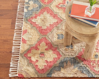 Könnte beinhalten: Ein gewebter Teppich mit einem sich wiederholenden Rautenmuster in Korallen-, Blau- und Beigetönen, der an der Kante mit einer weißen Franse versehen ist. Ein kleiner Holzhocker steht auf dem Teppich, darauf zwei gestapelte Bücher und eine kleine Glasvase.