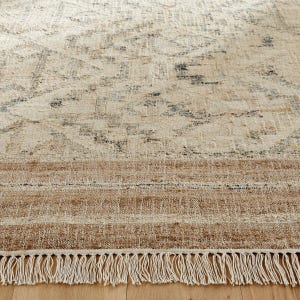 May include: A woven area rug featuring a cream-colored field with a faded charcoal gray geometric pattern. The rug includes a wide, textured border in a warm tan hue and is finished with a row of long, cream-colored fringe along the bottom edge.