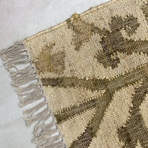 May include: Close-up of a beige and brown woven rug with a geometric pattern. The rug has a fringe on one side and is made of natural fibres. The design features diagonal lines and textured details.