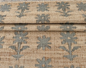 Könnte beinhalten: Ein gewebter Teppich mit einer naturfarbenen beigen Basis und einem sich wiederholenden Muster aus blaugrauen Blumenmotiven. Das Textil weist eine grobe Struktur und eine Fransenkante an der Oberseite auf, die das Design über die Stofffalten zeigt.