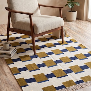 Puede incluir: Un sillón beige con brazos y patas de madera descansa sobre una alfombra rectangular. La alfombra presenta un patrón geométrico de cuadrados blancos, azul marino y verde oliva. La alfombra tiene un borde con flecos. Libros y una taza están sobre la alfombra.